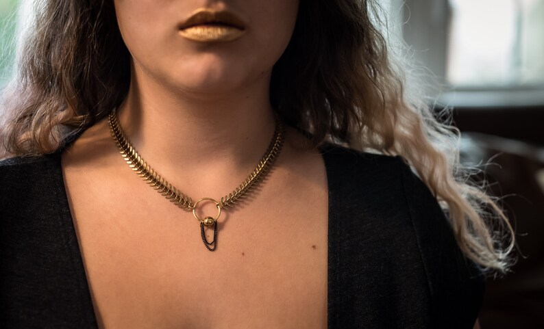May include: A gold chain choker necklace with a small round pendant and a black chain hanging from it.