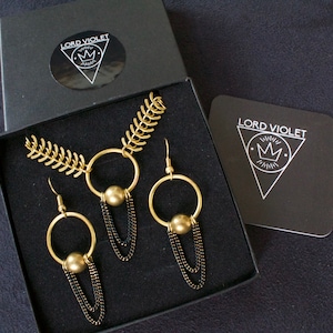 May include: A gold necklace and earrings set with black chain accents. The necklace features a large circle pendant with a gold ball. The earrings are hoop style with a gold ball hanging from the bottom. The jewelry is displayed in a black box with the brand name "LORD VIOLET" printed on the box.