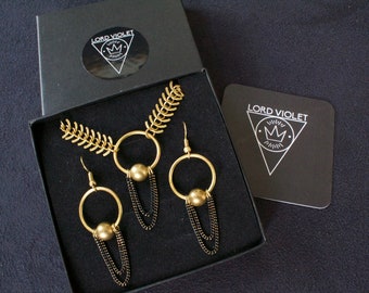 May include: A gold necklace and earrings set with black chain accents. The necklace features a large circle pendant with a gold ball. The earrings are hoop style with a gold ball hanging from the bottom. The jewelry is displayed in a black box with the brand name "LORD VIOLET" printed on the box.