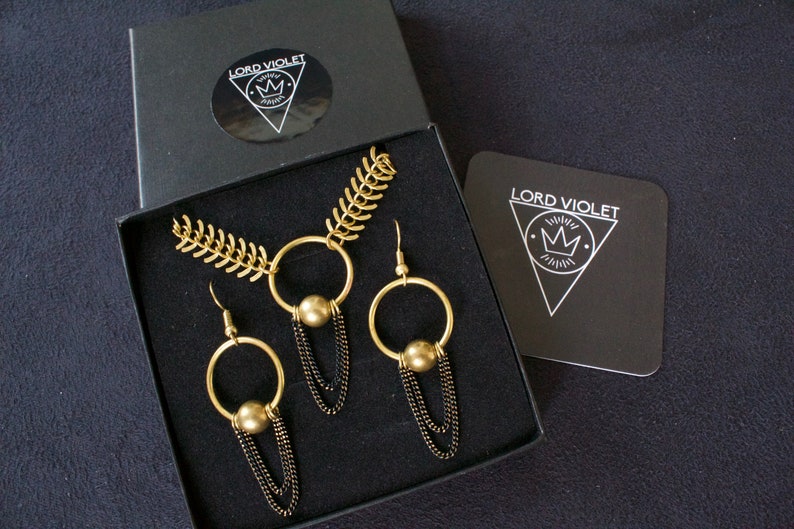 May include: A gold necklace and earrings set with black chain accents. The necklace features a large circle pendant with a gold ball. The earrings are hoop style with a gold ball hanging from the bottom. The jewelry is displayed in a black box with the brand name "LORD VIOLET" printed on the box.
