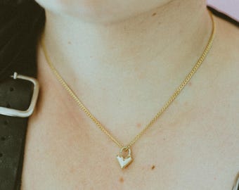 May include: A gold-colored necklace with a heart-shaped pendant. The pendant is a textured, light-colored metal. The necklace chain is a delicate, linked design. The background is a person's skin and a black garment.