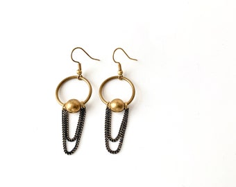 May include: A pair of brass-toned earrings with a circular design. Each earring features a small gold-colored ball suspended within a hoop, with two strands of dark gray chain hanging below. The earrings are displayed against a white background.