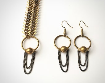 May include: A gold necklace with a chain made of interlocking triangles and a pendant with a circle and a dangling chain. Matching gold hoop earrings with a dangling chain.
