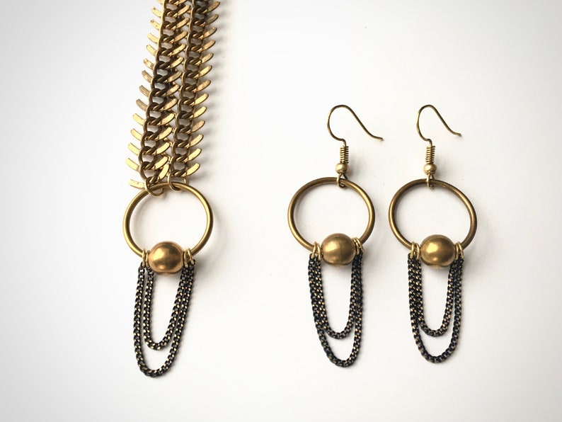 May include: A gold necklace with a chain made of interlocking triangles and a pendant with a circle and a gold bead. The necklace is paired with matching earrings with a circle and a gold bead, and three dangling chains.