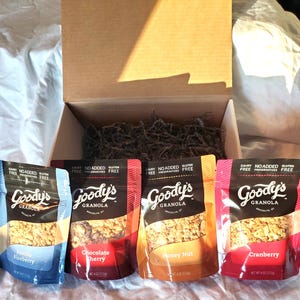 K&ouml;nnte beinhalten: Vier Beutel Goody's Granola in den Geschmacksrichtungen Vanille Blaubeere, Schoko Kirsche, Honig Nuss und Cranberry werden vor einer offenen Pappschachtel pr&auml;sentiert. Die Beutel sind in verschiedenen Farben gehalten und tragen die Aufschriften "Ohne Milchprodukte", "Ohne Zusatz von Konservierungsstoffen" und "Glutenfrei".