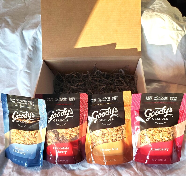 K&ouml;nnte beinhalten: Vier Beutel Goody's Granola in den Geschmacksrichtungen Vanille Blaubeere, Schoko Kirsche, Honig Nuss und Cranberry werden vor einer offenen Pappschachtel pr&auml;sentiert. Die Beutel sind in verschiedenen Farben gehalten und tragen die Aufschriften "Ohne Milchprodukte", "Ohne Zusatz von Konservierungsstoffen" und "Glutenfrei".