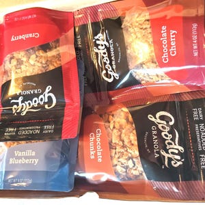K&ouml;nnte beinhalten: Eine Schachtel gef&uuml;llt mit Goody's Granola-Beuteln. Die Geschmacksrichtungen sind Cranberry, Chocolate Cherry, Vanilla Blueberry und Chocolate Chunks. Jeder Beutel wiegt 112g und ist milchfrei, ohne Zusatz von Konservierungsstoffen und glutenfrei.