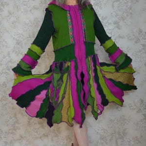 May include: A vibrant, handmade knit dress in shades of green, pink, and purple. The dress features a hooded top with a zipper, long sleeves, and a flared skirt. The sleeves and skirt have a layered, ruffled design. The model is wearing pink shoes.