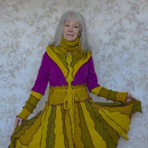 May include: A woman wearing a purple and yellow knitted dress with a long, flowing skirt. The dress has a unique design with a mix of textures and colours. She is wearing matching yellow shoes.