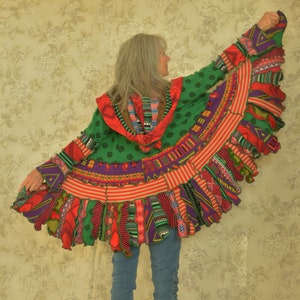 Könnte beinhalten: Ein grüner, roter und lila gestrickter Cardigan mit einem bunten Fransenbesatz. Der Cardigan wird über einer blauen Jeans getragen.