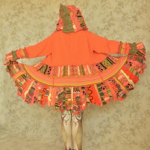 May include: A long, bright orange hooded cardigan with a colourful, multi-layered, crocheted trim. The cardigan has a loose fit and a flowing, layered hem.