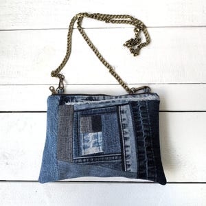 May include: A denim patchwork crossbody bag with a brass chain strap. The bag is made from various shades of blue denim, creating a unique geometric pattern. The zipper closure is visible at the top. The bag is approximately 10 inches wide.