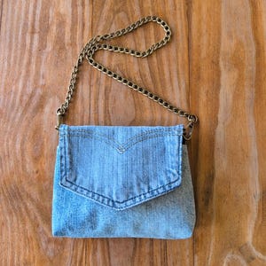 May include: A light blue denim crossbody bag with a gold-colored chain strap. The bag has a flap closure with decorative stitching. The bag is made of denim material and is designed to be worn over the shoulder.