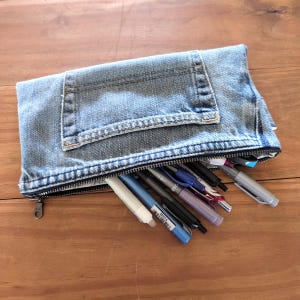 May include: A denim pencil case with a zipper closure and a pocket. The case is filled with various pens and pencils in shades of blue, black, and silver. The case is made from recycled denim.
