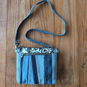 May include: A denim crossbody bag with a blue strap and a zipper closure. The bag's body is made from strips of blue denim in varying shades. The top features a black and white patterned fabric.