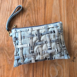 May include: A denim clutch with a woven design. The light blue bag features a zipper closure, a wrist strap, and a unique pattern created from strips of denim. The bag is approximately 10 inches wide and 7 inches tall.