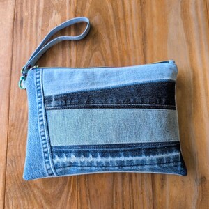 May include: A rectangular denim clutch with a wrist strap. The clutch is made from various shades of blue denim, with a zipper closure and a light blue strap. The design features horizontal stripes of different denim washes.