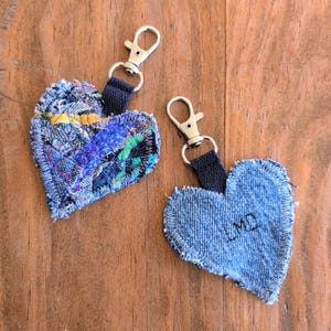 May include: Two denim heart-shaped keychains. One keychain is made of patchwork denim with colorful fabric accents. The other keychain is solid denim with the initials "LMD" embroidered in black thread.