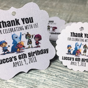 May include: Two white thank you tags with a cartoon image of a group of characters. The text on the tags reads "Thank You for celebrating with us! Lucca's 6th Birthday April 9, 2018".
