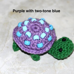 May include: A handmade crochet turtle in shades of purple and green. The turtle's shell is a vibrant purple, adorned with two-tone blue beads. The head and feet are green, with black bead eyes. The text "Purple with two-tone blue" is above the turtle.