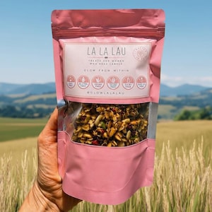 May include: A pink stand-up pouch of La La Lau granola with a clear window showing a mix of nuts, seeds, and dried fruit. The label features the text "La La Lau," "Treats for women who read labels," "Glow from within," and six circular dietary icons.