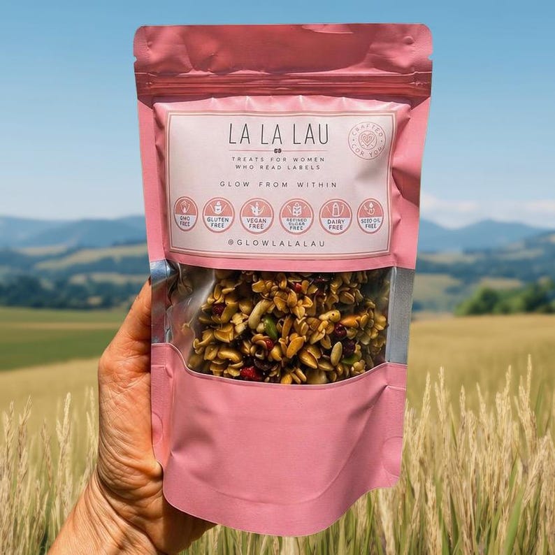 May include: A pink stand-up pouch of La La Lau granola with a clear window showing a mix of nuts, seeds, and dried fruit. The label features the text "La La Lau," "Treats for women who read labels," "Glow from within," and six circular dietary icons.