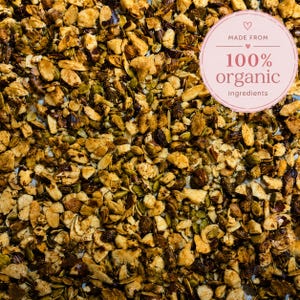 May include: A mixture of toasted nuts and green pumpkin seeds with a circular pink label in the upper right corner that reads MADE FROM 100% organic ingredients with a small heart icon above and below the text in a light pink font.