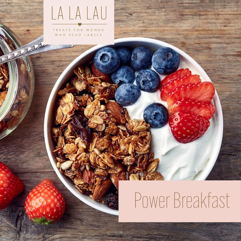 May include: A white bowl filled with granola, yogurt, fresh blueberries, and sliced strawberries. The text LA LA LAU TREATS FOR WOMEN WHO READ LABELS and Power Breakfast is visible. A glass jar with granola and a metal spoon are also present.