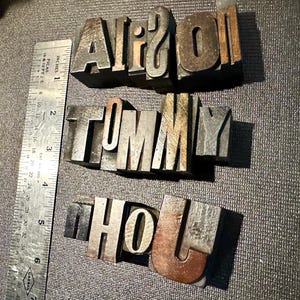May include: Three rows of vintage metal letterpress blocks spelling out the words AIRSON, TOMMY, and HOLJ. A metal ruler is positioned vertically on the left side, showing the blocks are approximately 1.5 inches in height.