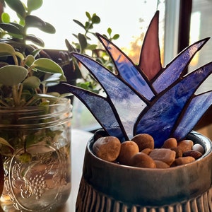 May include: A stained glass succulent in a blue ceramic pot with small rocks. The succulent is made of blue, purple, and pink glass.