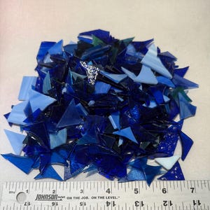 May include: A pile of blue and light blue glass mosaic tiles in various sizes and shapes. The tiles are arranged in a random pattern and are ready to be used in a craft project.