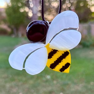May include: A handcrafted bee pendant with a black cord. The bee features white wings, a yellow body with black stripes, and a dark brown head. The pendant is likely made of glass or a similar material.