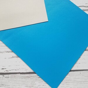 May include: A sheet of bright blue paper with a white sheet of paper partially overlapping it. The blue paper has a subtle textured surface.