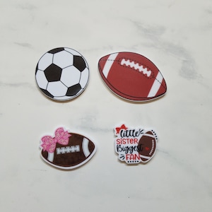 May include: Four iron-on patches featuring sports themes. One patch is a white and black soccer ball. Another patch is a brown and white football. The third patch is a brown and white football with a pink bow and the text "little sister biggest fan". The fourth patch is a brown and white football with the text "little sister biggest fan".