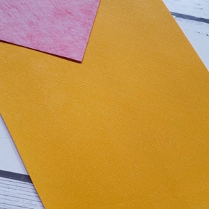 May include: A close-up of a textured yellow sheet of craft paper. The paper is slightly textured and has a subtle sheen. The paper is lying on a white surface.