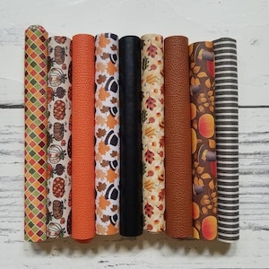 May include: Eight rolls of patterned faux leather in various fall-themed designs, including pumpkins, pinecones, leaves, and plaid. The rolls are arranged horizontally on a white wooden surface.