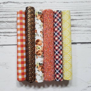 May include: Six rolls of patterned faux leather in various colors and designs, including orange, red, white, yellow, and brown. The rolls are arranged horizontally on a white wooden surface.