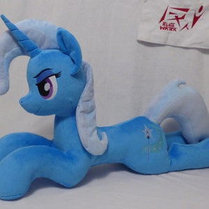 May include: A plush, light blue pony with a curved horn and flowing mane. The pony has a crescent moon and star design on its flank. The eyes are purple and black, with long eyelashes. The pony is lying down.