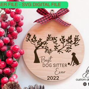 Può includere: Un ornamento natalizio in legno con una silhouette marrone di un cane seduto sotto un albero con foglie. L'ornamento recita "Best Dog Sitter Ever 2022" ed è decorato con un nastro rosso e nero a scacchi.