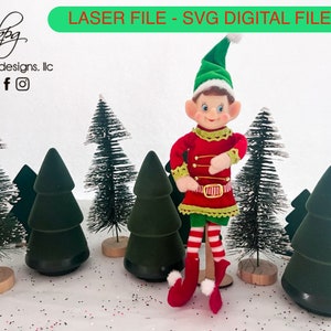 Può includere: Un pupazzo di elfo rosso e verde con un bordo di pelliccia bianca si trova di fronte a cinque alberi di Natale verdi in miniatura. L'elfo indossa una cintura a righe rosso e bianco e calze a righe rosso e bianco. Gli alberi sono su una superficie bianca con glitter rosso e bianco.