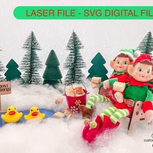 May include: Two elves sitting on a wooden bench with a red and green striped blanket. They are holding small pieces of bread. The bench is decorated with snowflakes. There is a sign that says "Please don't feed the ducks!" and two yellow rubber ducks in a small pond. The scene is set in a snowy forest with small green pine trees.