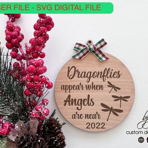 May include: A round wooden ornament with the text "Dragonflies appear when Angels are near 2022" and three dragonfly illustrations. The ornament is decorated with a red and white plaid ribbon.