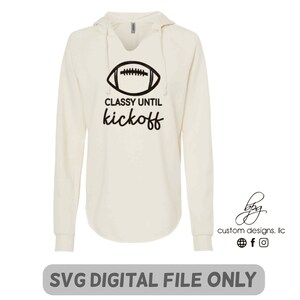 May include: Cream-coloured hoodie with a football graphic and the text "Classy until kickoff." The long-sleeved shirt has a v-neck and a hood. The bottom of the shirt has a curved hemline. The words "SVG DIGITAL FILE ONLY" are at the bottom.
