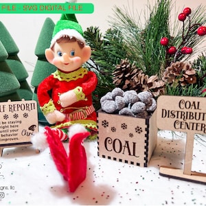 Puede incluir: Una escena navideña con una figura de elfo, pequeños árboles verdes y letreros. Un letrero dice "Elf Report" con un mensaje sobre el comportamiento. Otro letrero dice "Coal Distribution Center". Una caja de madera contiene carbón.