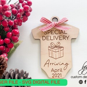 May include: A wooden baby onesie ornament with the text "Special Delivery" and "Arriving April 2021". The ornament is decorated with a red and white striped ribbon and a drawing of a gift box with a bow.