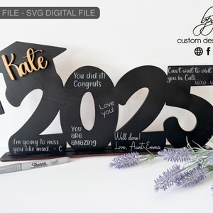 Op de afbeelding: Een zwart houten 2023 afstudeerbord met een afstudeerpet er bovenop. Het bord is versierd met gouden letters en heeft ruimte voor berichten. Het bord staat op een tafel met een zilveren Sharpie-marker.