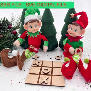 May include: Two elf dolls playing a game of noughts and crosses on a wooden board. The elves are wearing red and green elf hats and outfits. The board is surrounded by green pine trees and white glitter.