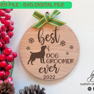 May include: A round wooden ornament with the text "best dog groomer ever" and the year "2022". The ornament is decorated with a small dog silhouette and snowflakes. A green and white ribbon is attached to the top of the ornament.