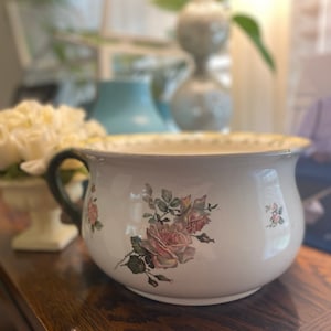 May include: A white ceramic chamber pot with a black handle and a floral design featuring pink roses and green leaves.
