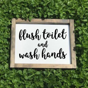 May include: A white wooden sign with black text that reads "flush toilet and wash hands". The sign is sitting on a bed of green plants.